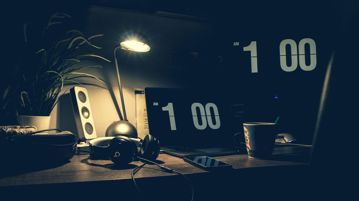 A dimly lit modern office desk with a laptop, headphones, and speakers at 1:00 AM.